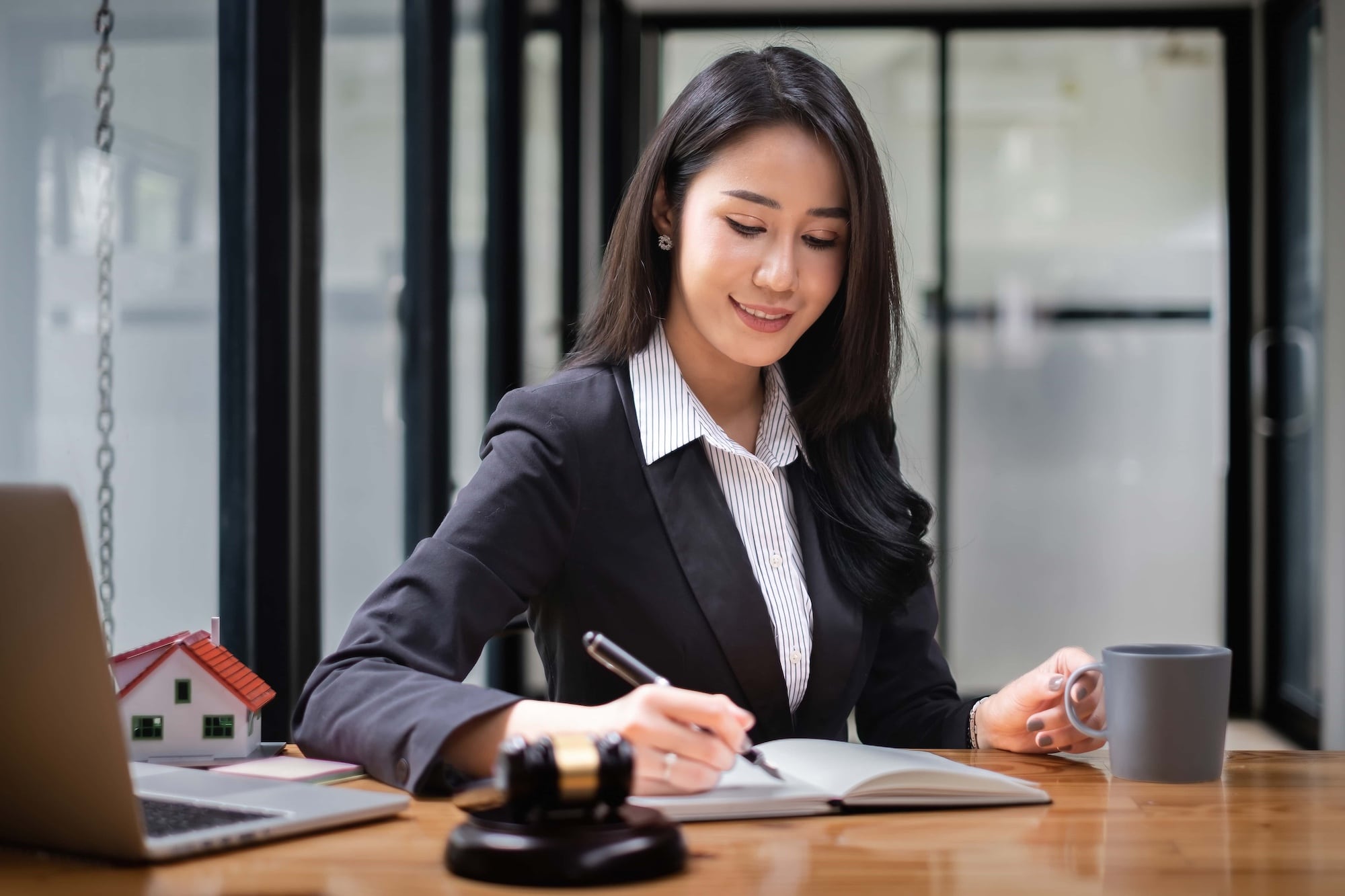 A woman in a business suit writes in a notebook at a desk with a gavel, a model house, a laptop, and a coffee mug. She is in a modern office setting and appears focused and content.