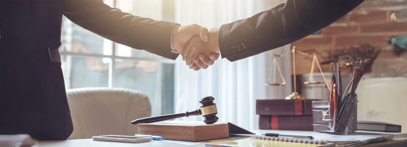 Two people in suits are shaking hands over a desk with a gavel, a stack of books, and scales of justice. The scene is set in a well-lit office, suggesting a legal or business agreement.
