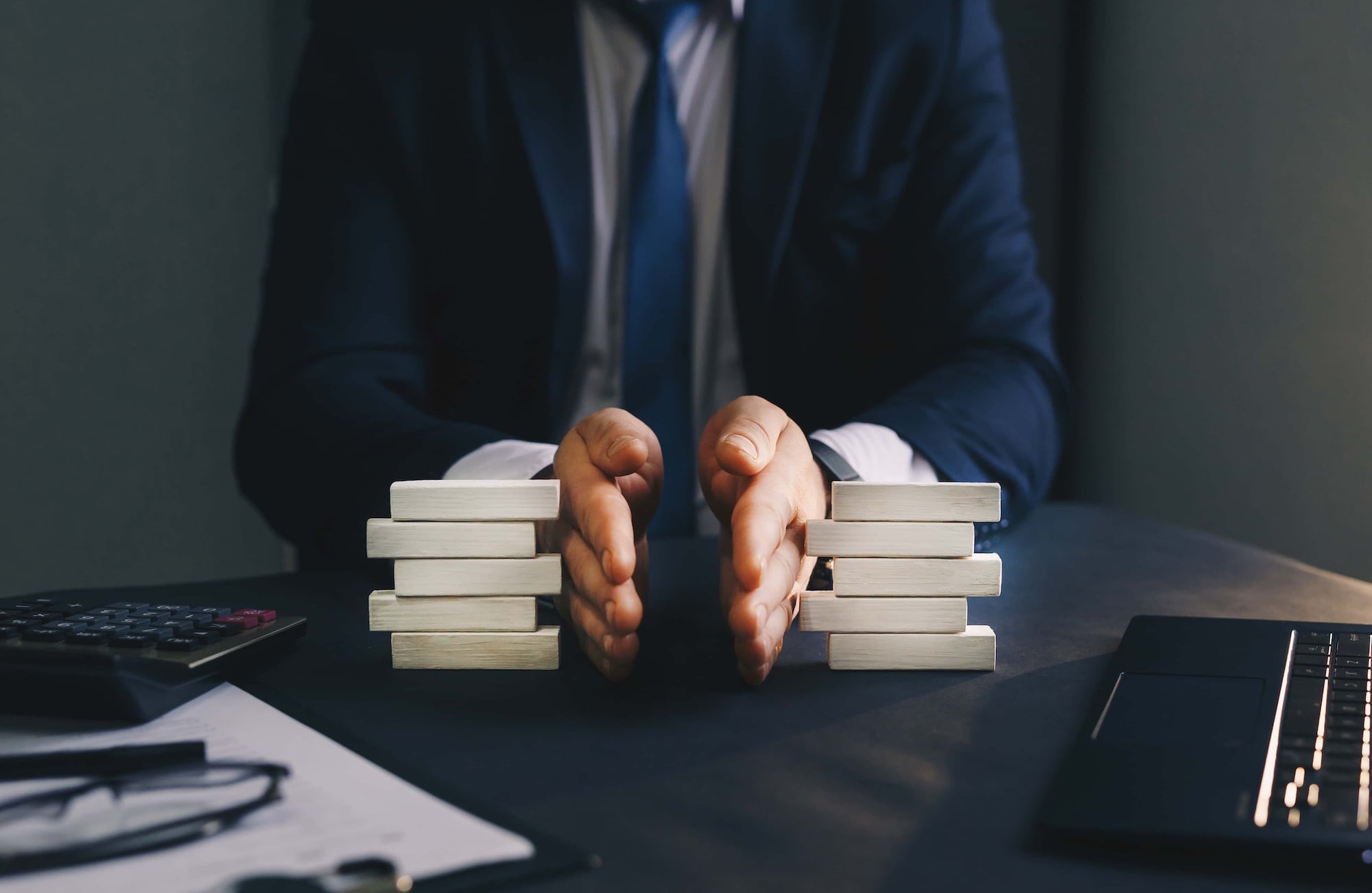 A person in a suit sits at a desk with hands separating two stacks of wooden blocks. A laptop, calculator, and papers are also on the desk. The scene suggests negotiation or decision-making.