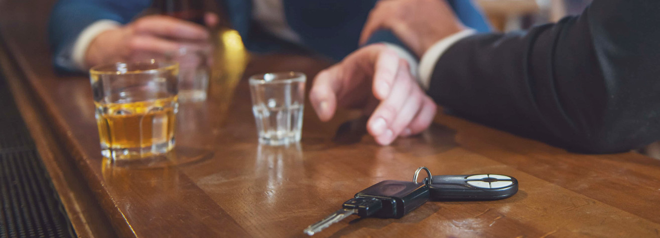 Two people sitting at a bar counter, each with a drink in front of them. A set of car keys is placed on the counter near their hands, suggesting a concern about drinking and driving.