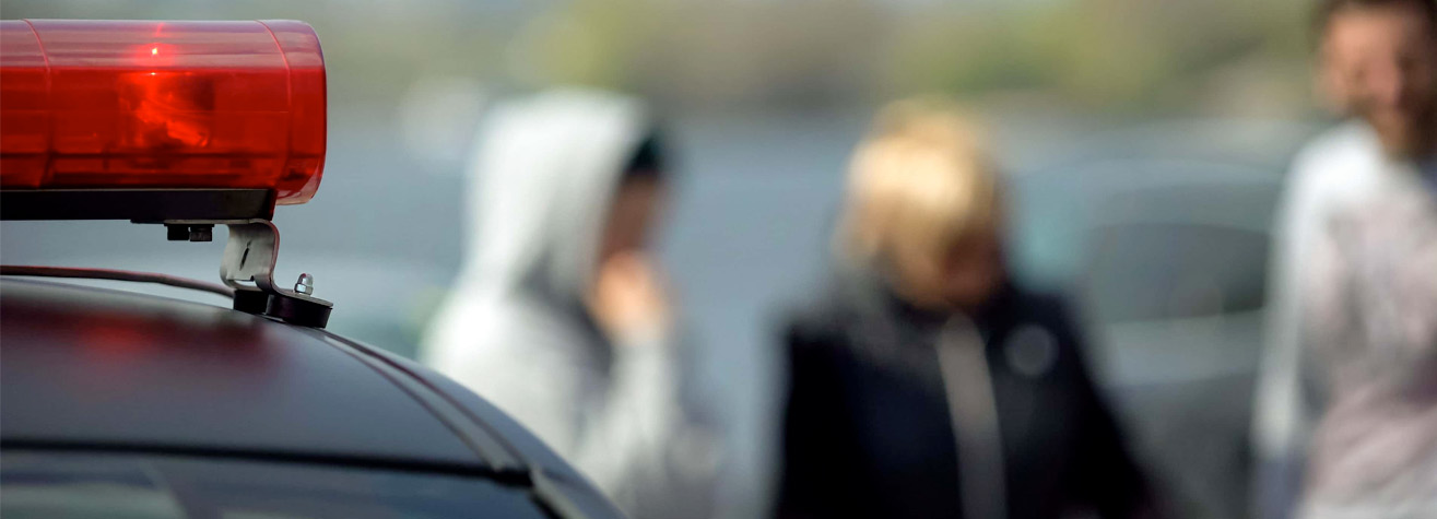 A red police siren on top of a vehicle in the foreground. In the blurred background, three people are standing, with one wearing a hoodie and the others in casual jackets.
