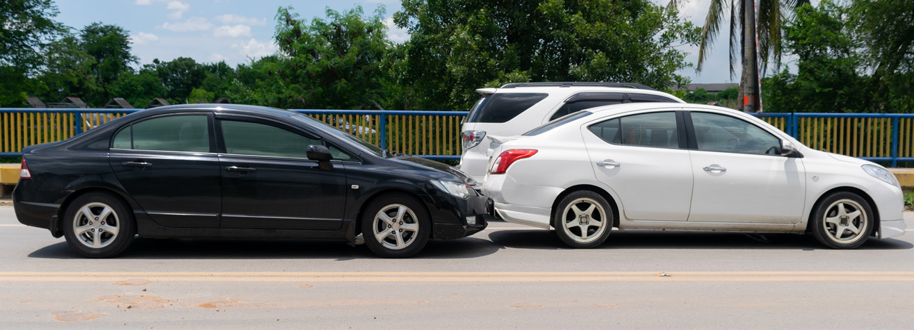 A black sedan and a white sedan are parked closely in a line on a paved road. A white SUV is parked in front of them. The background features trees and a blue and yellow fence under a partly cloudy sky.