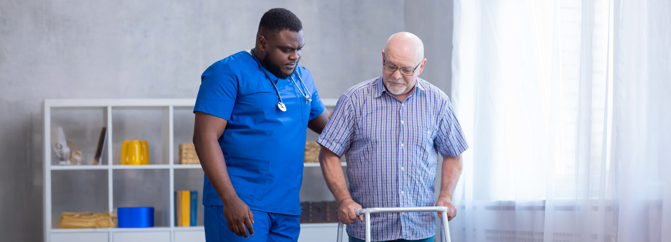 A healthcare professional in blue scrubs assists an elderly man using a walker. They are in a well-lit room with a white shelving unit and decorative items in the background. The elderly man wears glasses and a plaid shirt.