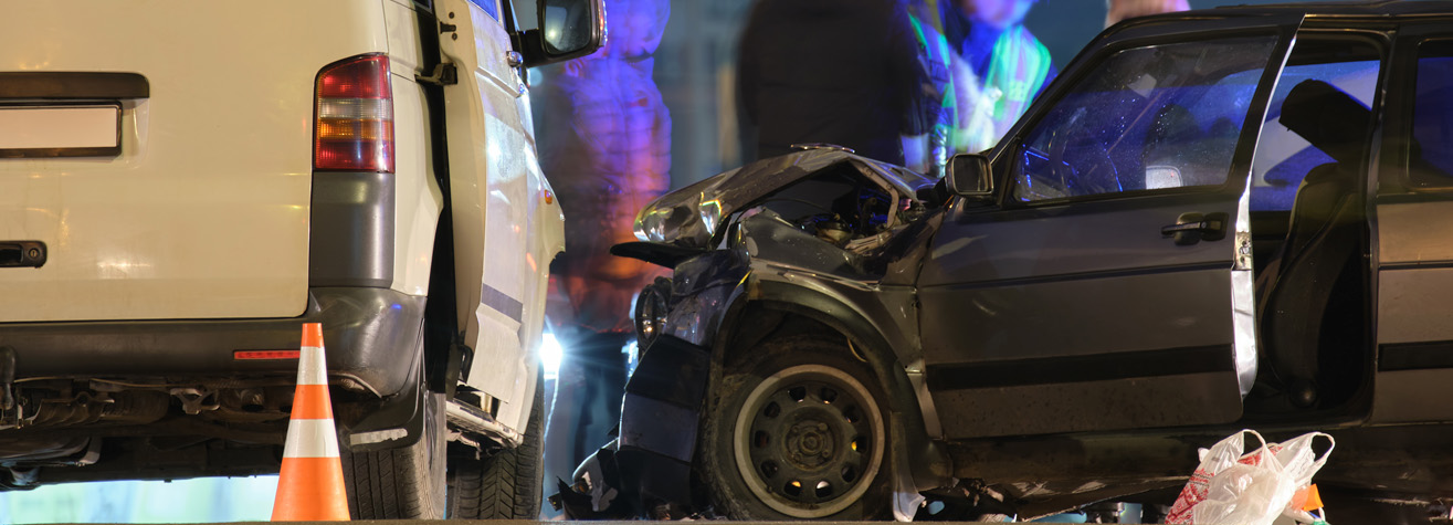 A car crash scene at night involving a white van and a black car with a damaged front. Orange traffic cones are placed nearby, and people in reflective vests are in the background, with a shopping bag visible on the ground.