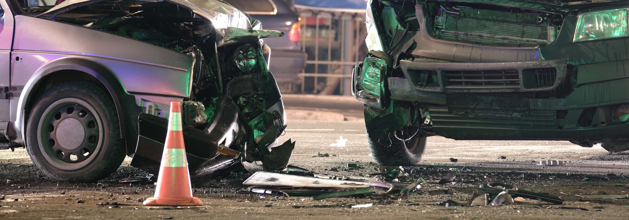 Two heavily damaged cars in a front-end collision on a street, surrounded by debris. A traffic cone is in the foreground, and a blurred barrier is visible in the background, indicating an accident scene.