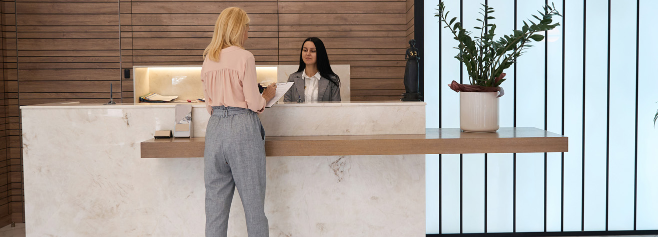 A woman in a pink blouse stands at a marble reception desk, facing another woman in a black blazer sitting behind the desk. A large potted plant decorates the counter. The background features wooden paneling and vertical window blinds.