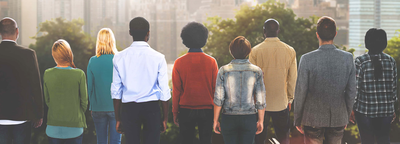 A diverse group of people stand outdoors with their backs to the camera, looking at a city skyline. The group includes individuals of various races and genders, wearing casual clothing. Trees and buildings are visible in the background.