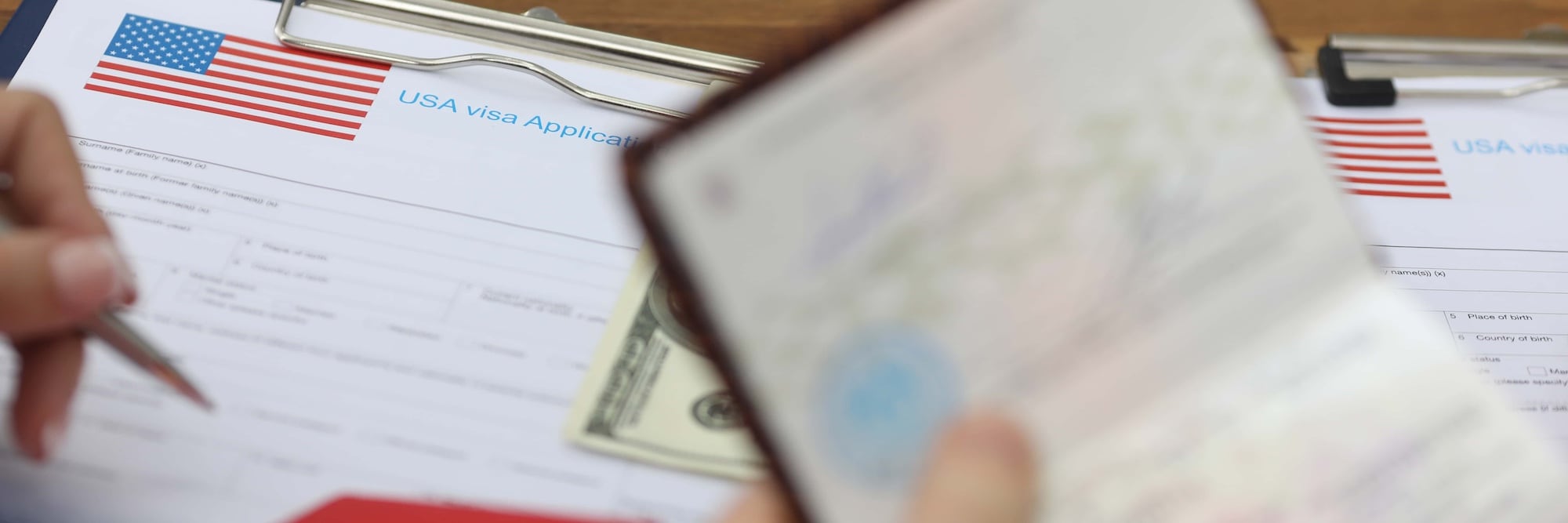 A person fills out a USA visa application form on a clipboard. A hundred-dollar bill is visible nearby, and another hand holds a passport. The form and the passport are in focus, emphasizing travel-related paperwork.
