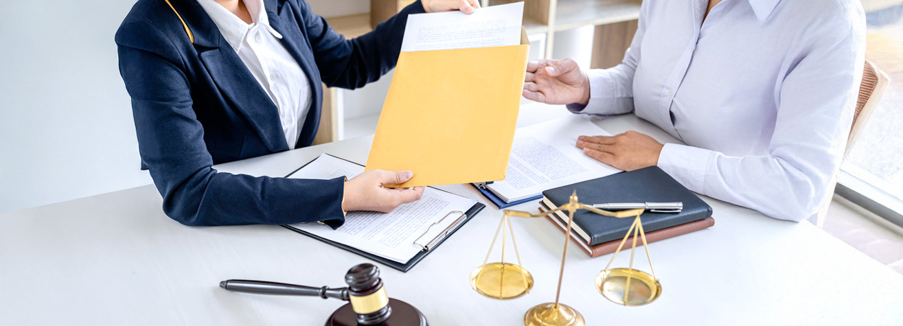 Two people at a desk with documents. One person hands a folder to the other. A gavel and scales of justice are on the table, suggesting a legal setting.