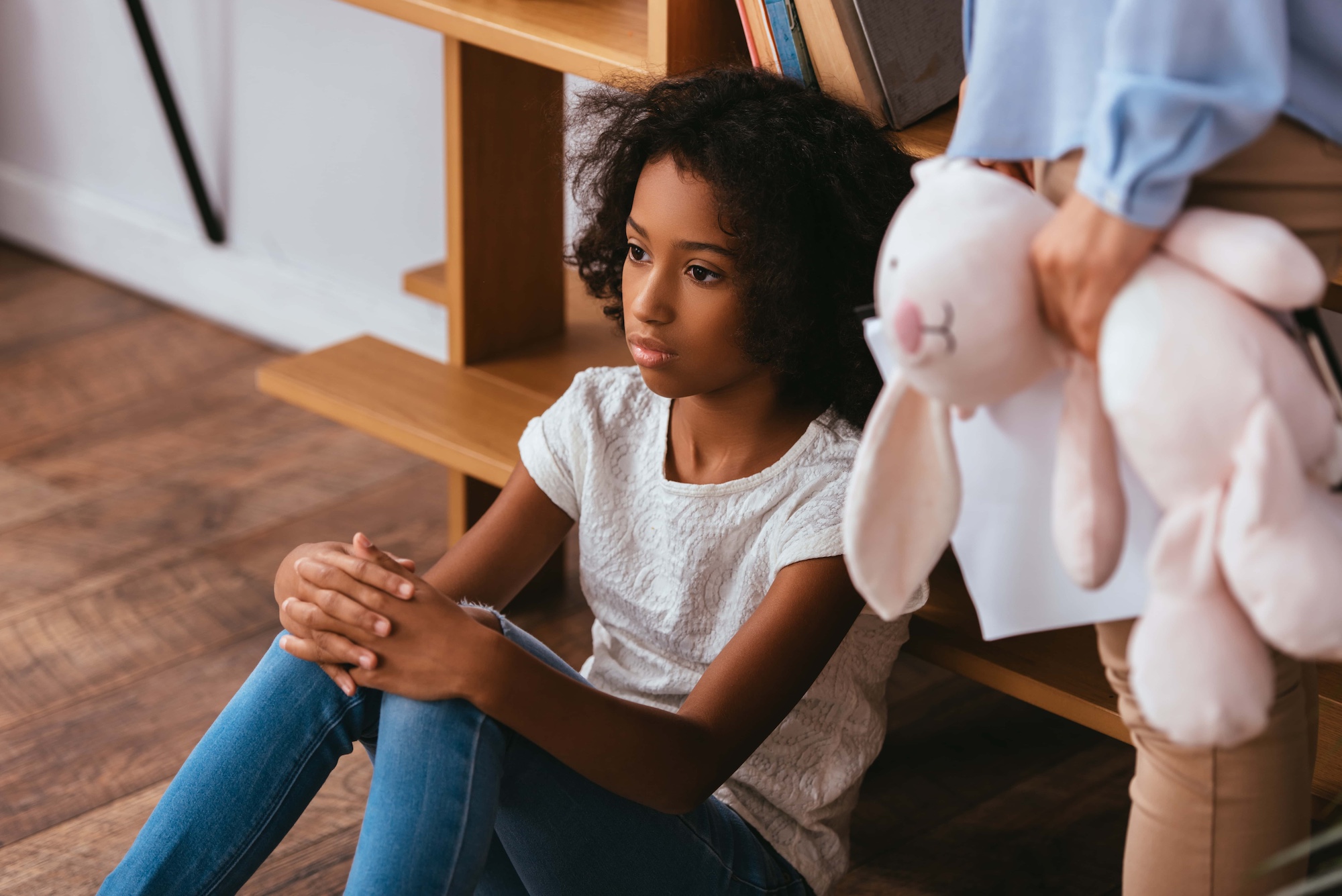 A young girl with curly hair sits on the floor looking thoughtful, clasping her knees. Beside her, an adult seen from the waist down holds a stuffed rabbit and papers. The setting appears to be a room with wooden flooring and bookshelves.
