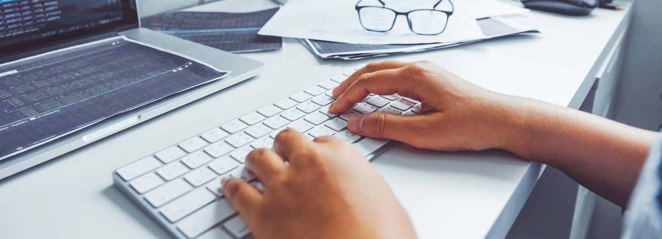 A person is typing on a white keyboard in front of a laptop. Papers and glasses are scattered on the desk, alongside a credit card. The laptop screen displays lines of code.