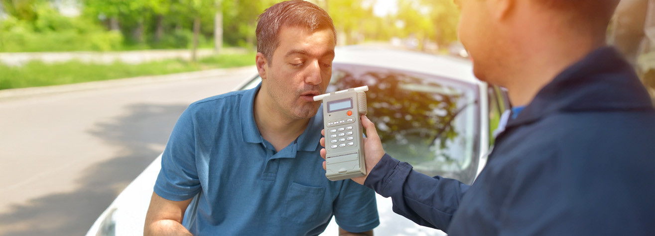 A man in a blue shirt blows into a breathalyzer held by another person standing in front of him. They are outdoors near a car, with trees and a road visible in the background.