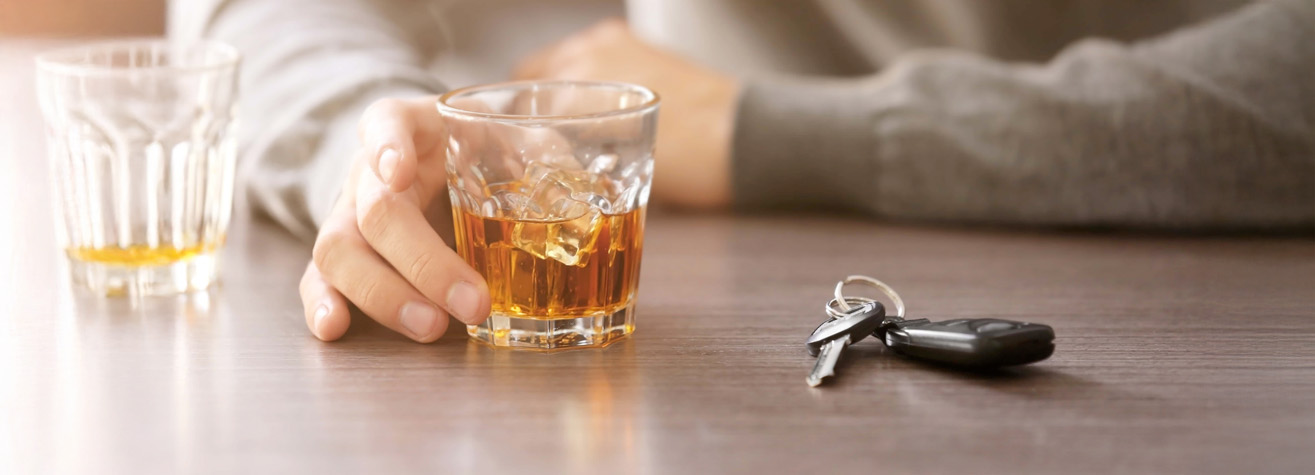 A person in a gray sweater holds a glass of whiskey on a wooden table. Car keys are placed nearby, suggesting the theme of drinking and driving. The scene is softly lit, emphasizing the contrast between the drink and the keys.