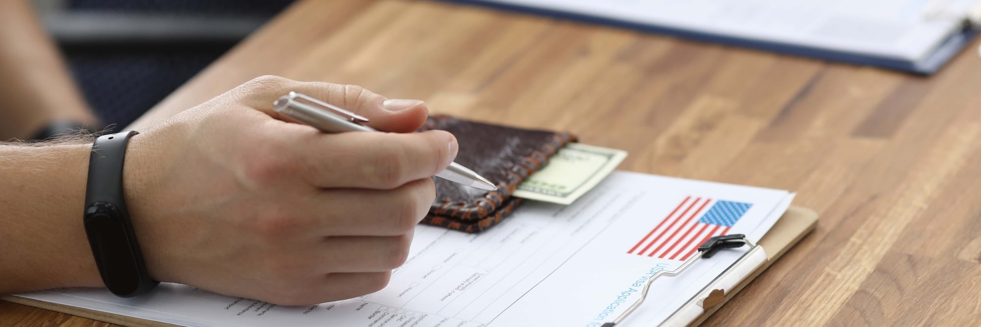A person is filling out a form on a clipboard with a small American flag in the corner. They hold a pen in their right hand and wear a black wristband. A wallet containing cash sits on the table.