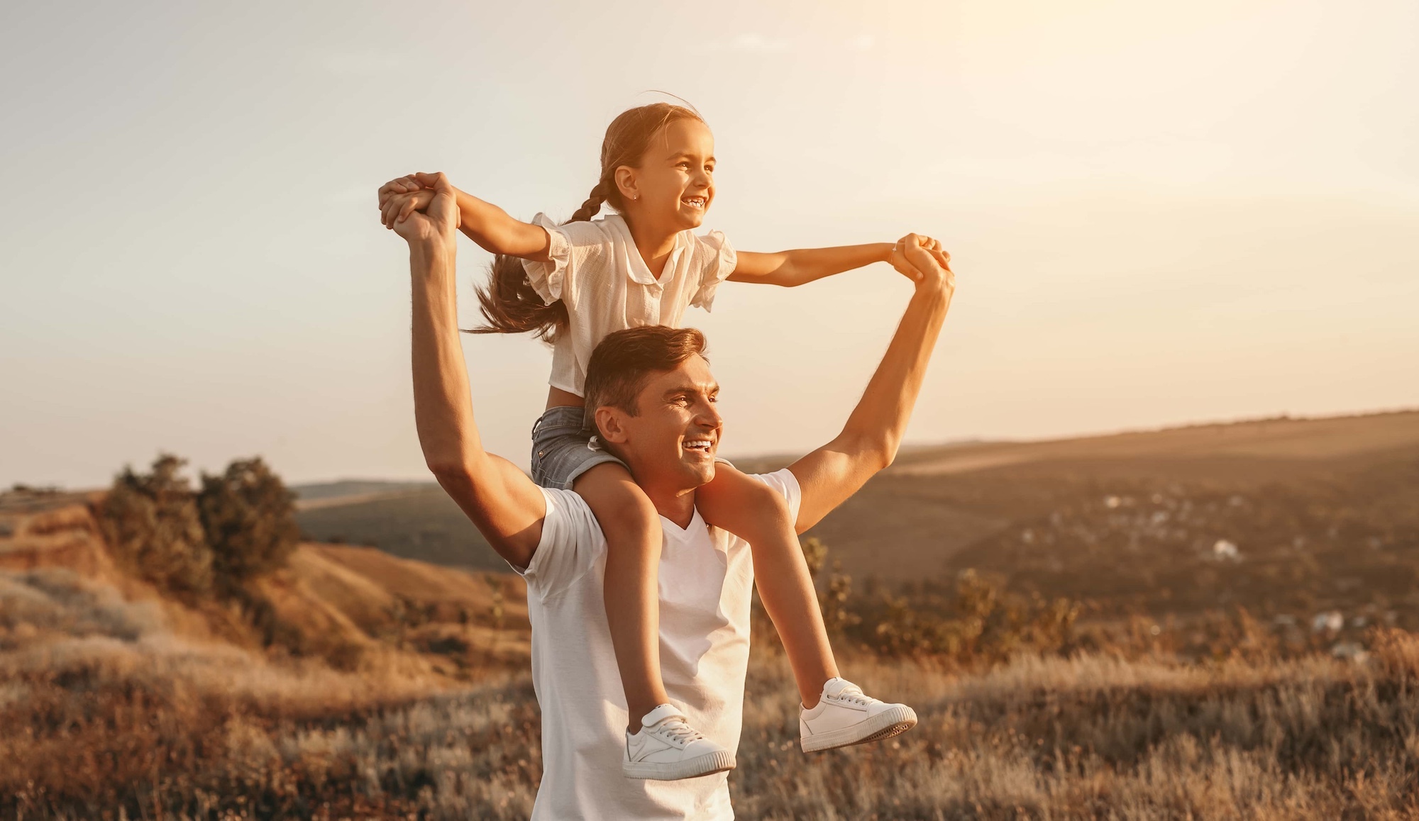 A man is carrying a smiling child on his shoulders in a grassy, sunlit landscape. The child has her arms outstretched, and both are looking ahead with content expressions. The background features rolling hills and a warm, golden sky.