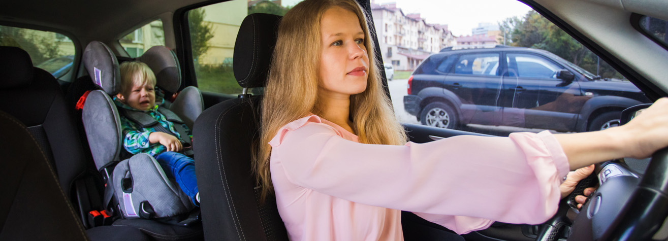 A woman with long blonde hair is driving a car. In the back seat, a young child in a car seat appears upset and is crying. The scene is set in an urban area with other cars and buildings visible outside.