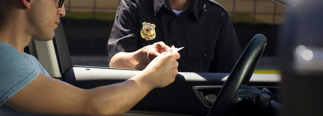 A driver in a car is handing a document to a police officer standing outside. The officer is wearing a uniform with a badge. The driver's window is open.