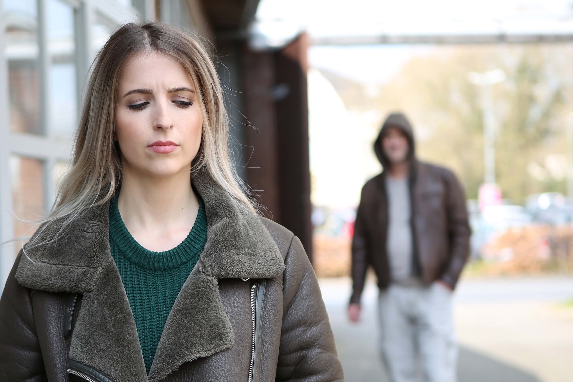 A woman with long blonde hair, wearing a green sweater and brown jacket, walks forward looking concerned. Behind her, a blurry figure in a hooded jacket and light pants appears to be following her. The background is an outdoor area.