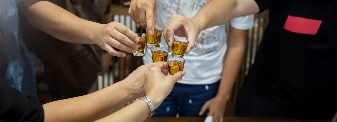 A group of people clinking shot glasses filled with a golden liquid in a celebratory gesture. The focus is on their hands and the glasses, with various shirts visible in the background.