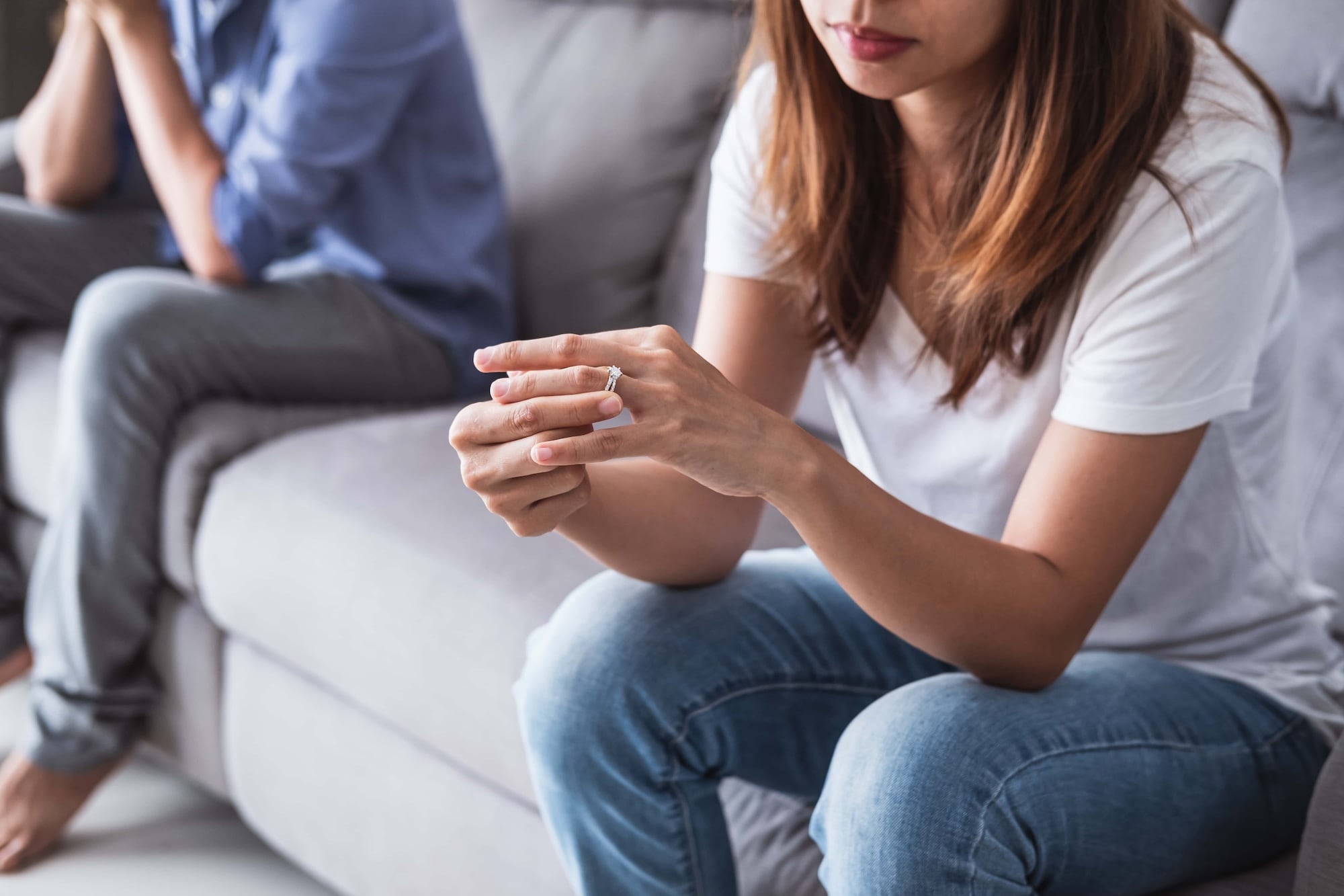 A woman in a white t-shirt and jeans sits on a couch, looking down at a ring in her hands. A man in a blue shirt sits at a distance in the background, slightly blurred. The scene suggests a tense or contemplative moment.