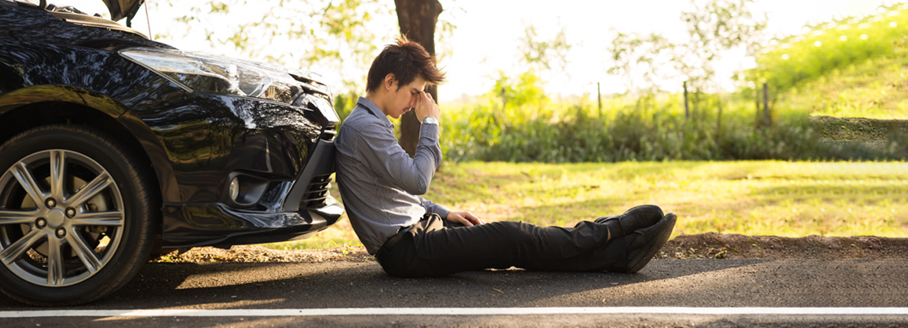 A man in a blue shirt and black pants sits on the roadside, leaning against the front bumper of a car with the hood open. He appears stressed, resting his head on his hand. The background features grass and trees in soft daylight.