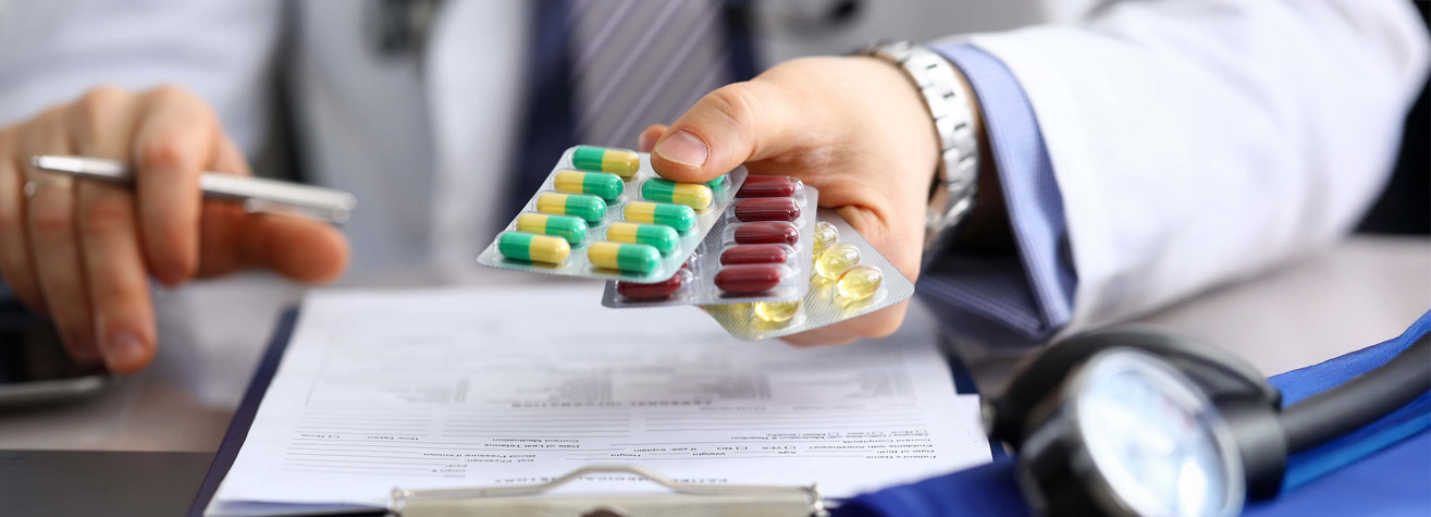 A doctor in a white coat extends a blister pack of multicolored pills over a document on a clipboard. A stethoscope is visible in the foreground. The doctor holds a pen in the other hand, suggesting a consultation setting.