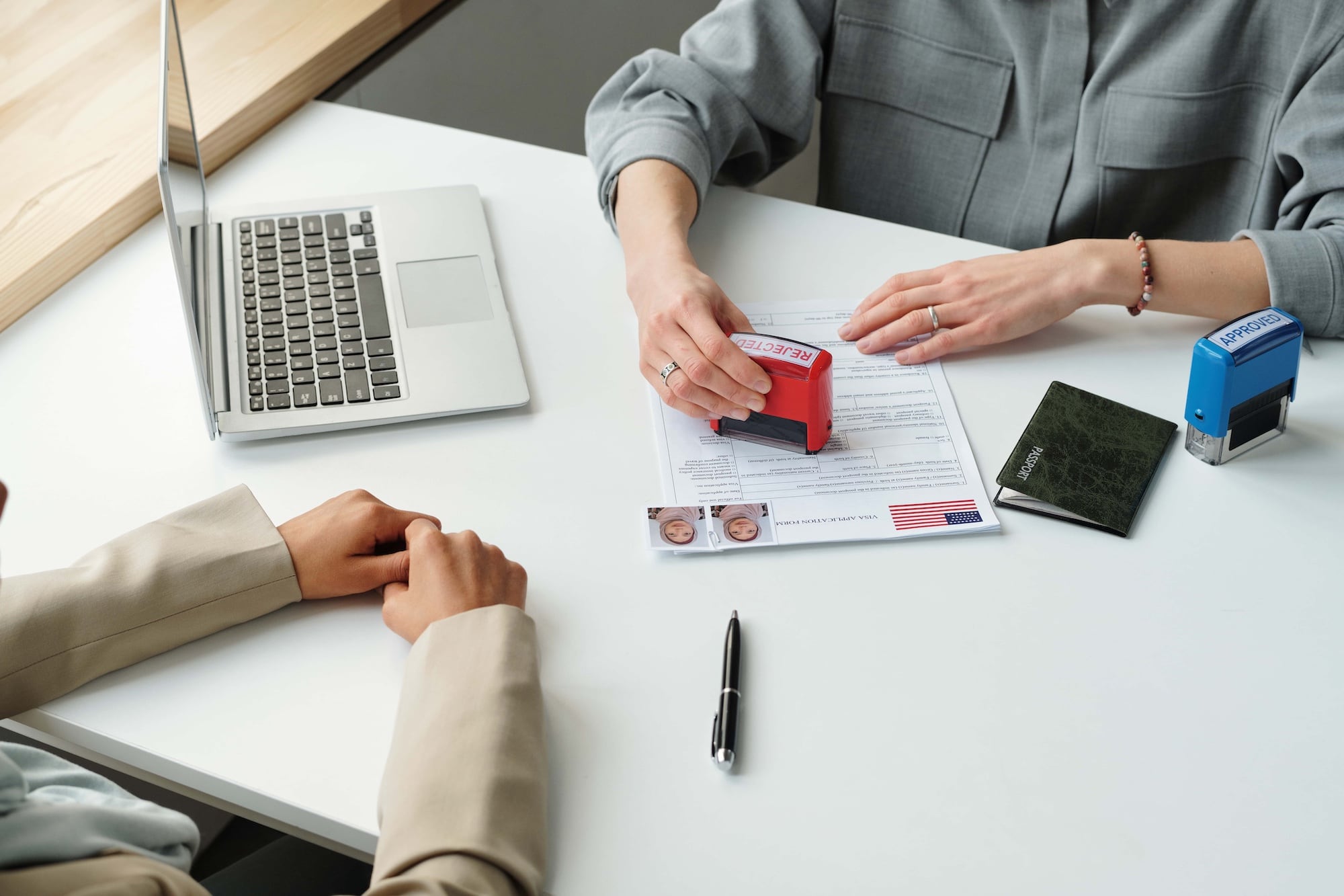 Two people sit at a desk. One person stamps a document near a laptop, while the other sits with hands folded. A pen and green passport are on the table. The document features photos and a small U.S. flag.