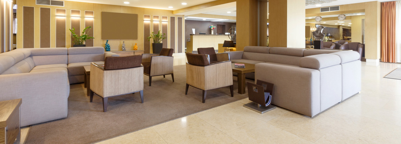A modern hotel lobby with beige sofas and wooden armchairs arranged around a rug. Decorative vases are displayed on a shelf, and a reception desk is visible in the background. The area is well-lit with recessed ceiling lights.