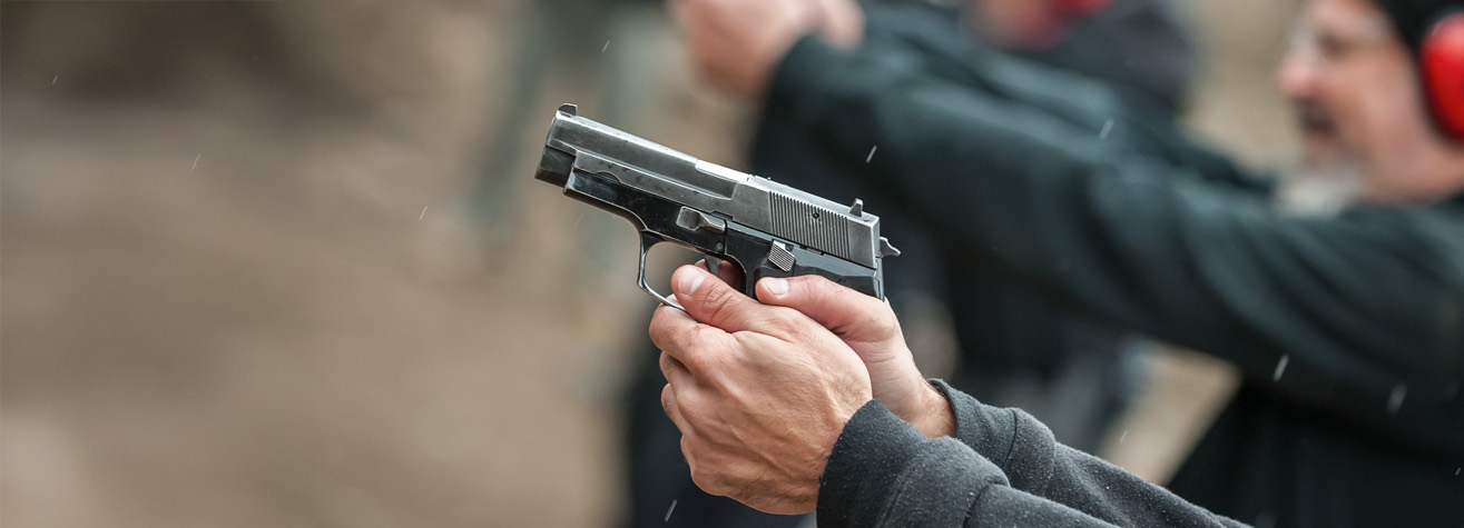 Close-up of a person's hands holding a silver handgun. In the background, more people are visible in a blurred view, each aiming a firearm. The scene suggests a shooting range, with focus on the weapon.