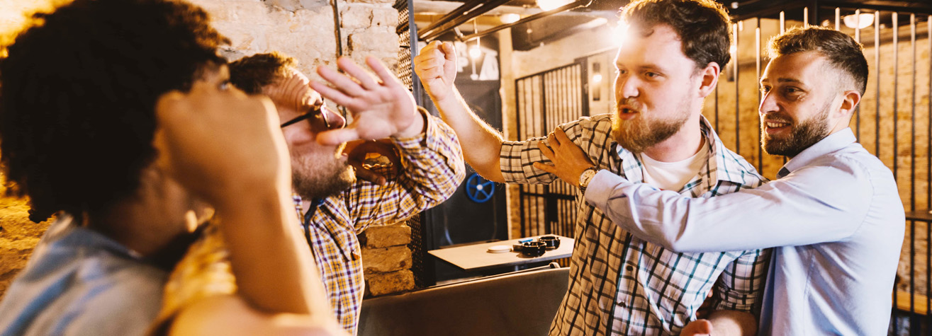 Four men in a pub appear engaged in a heated argument. Two men are raising their hands defensively, while the other two try to restrain each other. The scene is tense, set against a background of exposed brick walls and tables.