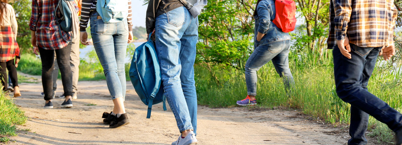 A group of people walking on a dirt path surrounded by greenery. They wear casual clothing and carry backpacks, suggesting a hiking or outdoor activity. The scene is bright, indicating daytime.