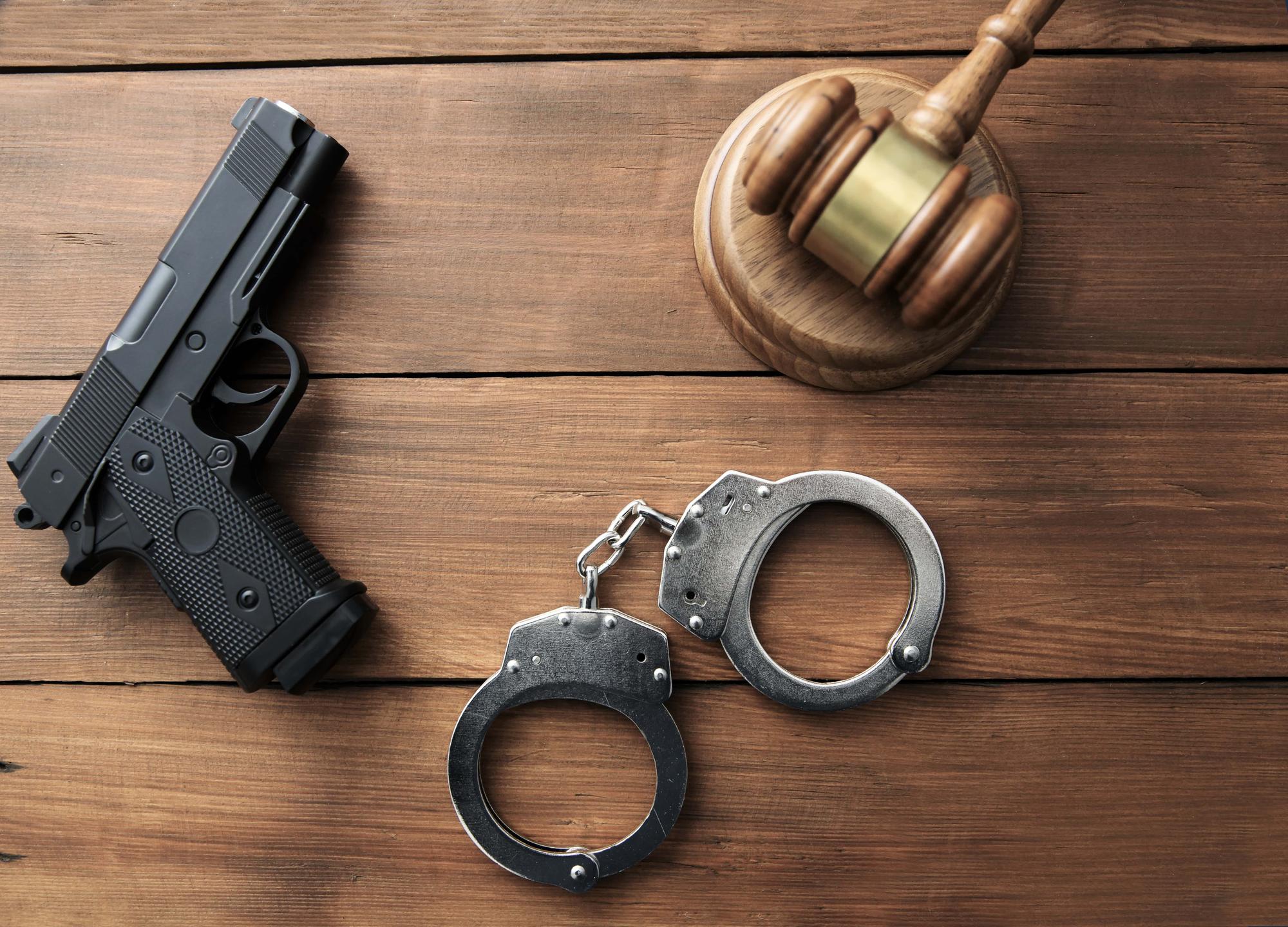 A black handgun, metal handcuffs, and a wooden gavel are arranged on a wooden surface. The gavel rests on a round block, symbolizing law and justice.