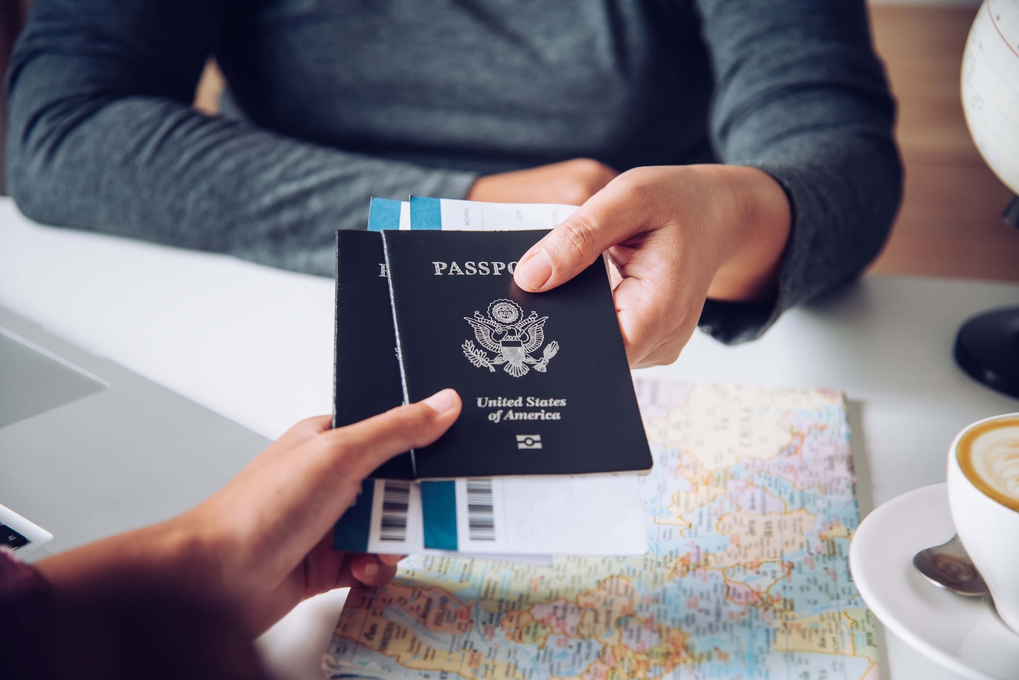 Close-up of two people exchanging a U.S. passport and boarding pass over a table with a world map. A laptop and a cappuccino cup are partially visible, suggesting a travel planning scene.