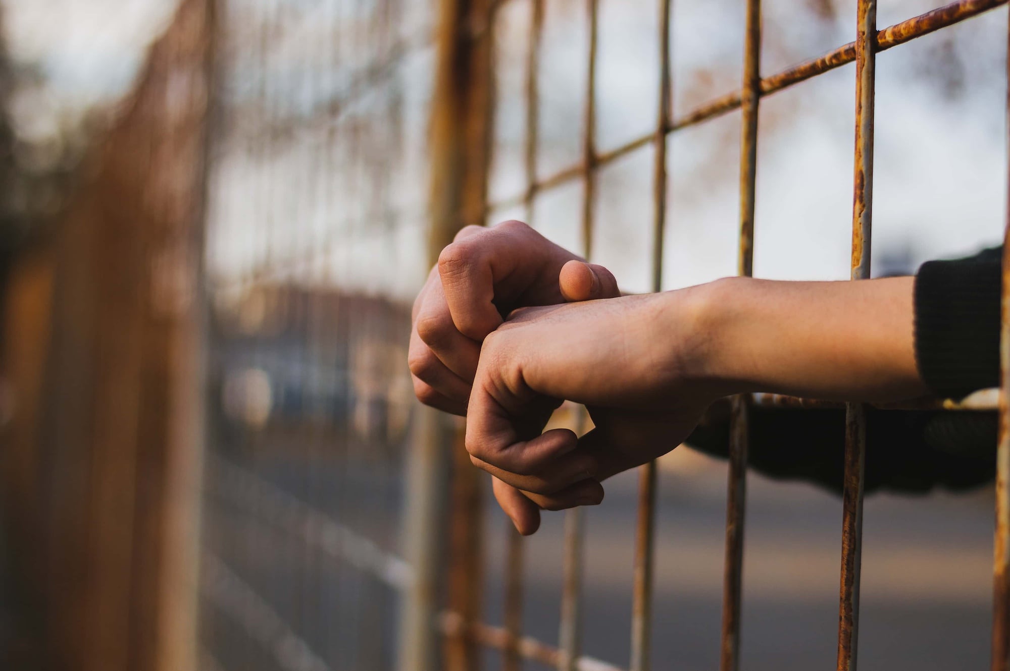 Close-up of hands resting on a metal fence, with fingers interlocked. The background is blurred, showing an outdoor setting in soft focus. The lighting suggests a late afternoon or early evening atmosphere.