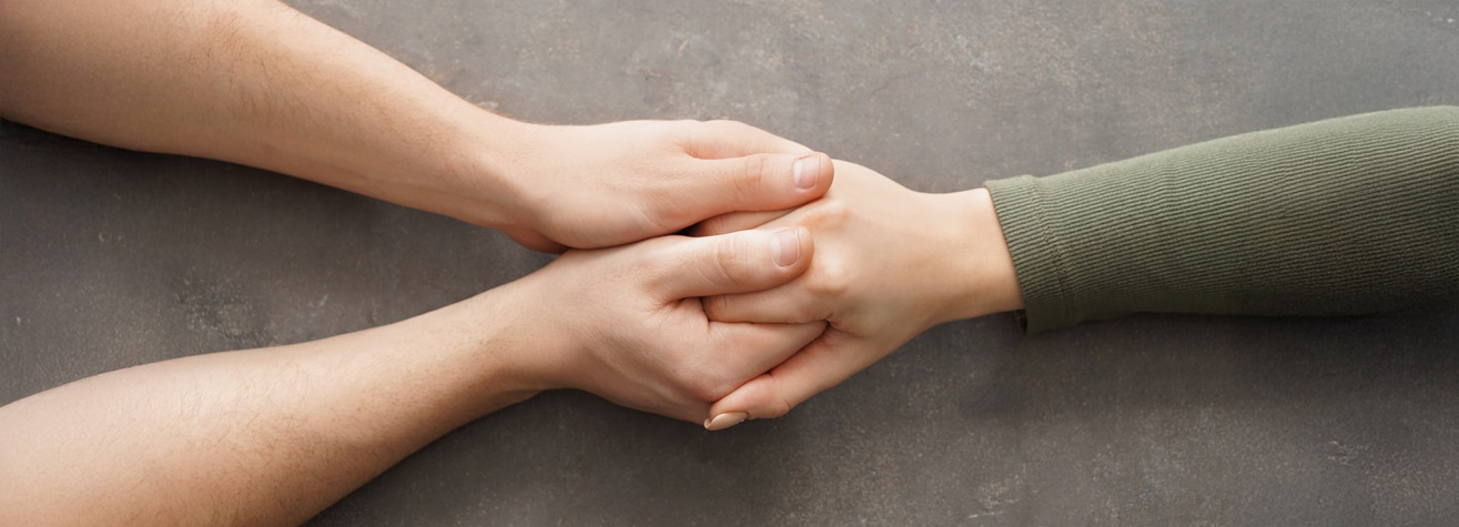 Two hands are gently holding another hand over a gray surface. One person is wearing a long-sleeved green shirt, while the other person has bare arms, suggesting a comforting or supportive gesture.