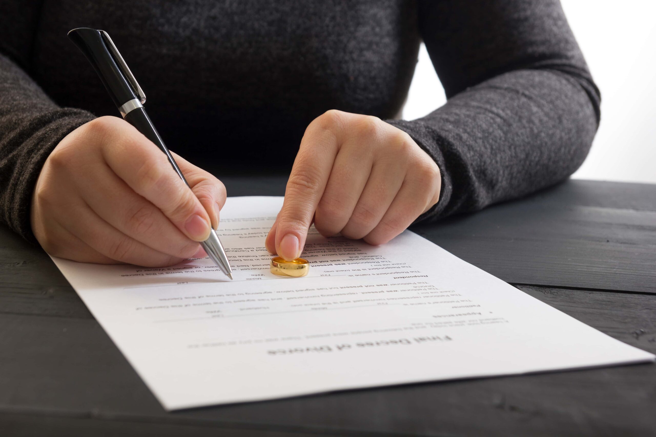 A person in a gray sweater signs a document with a pen on a dark wooden table. Their other hand rests on the paper, holding a gold ring. The scene suggests signing a formal or legal document.