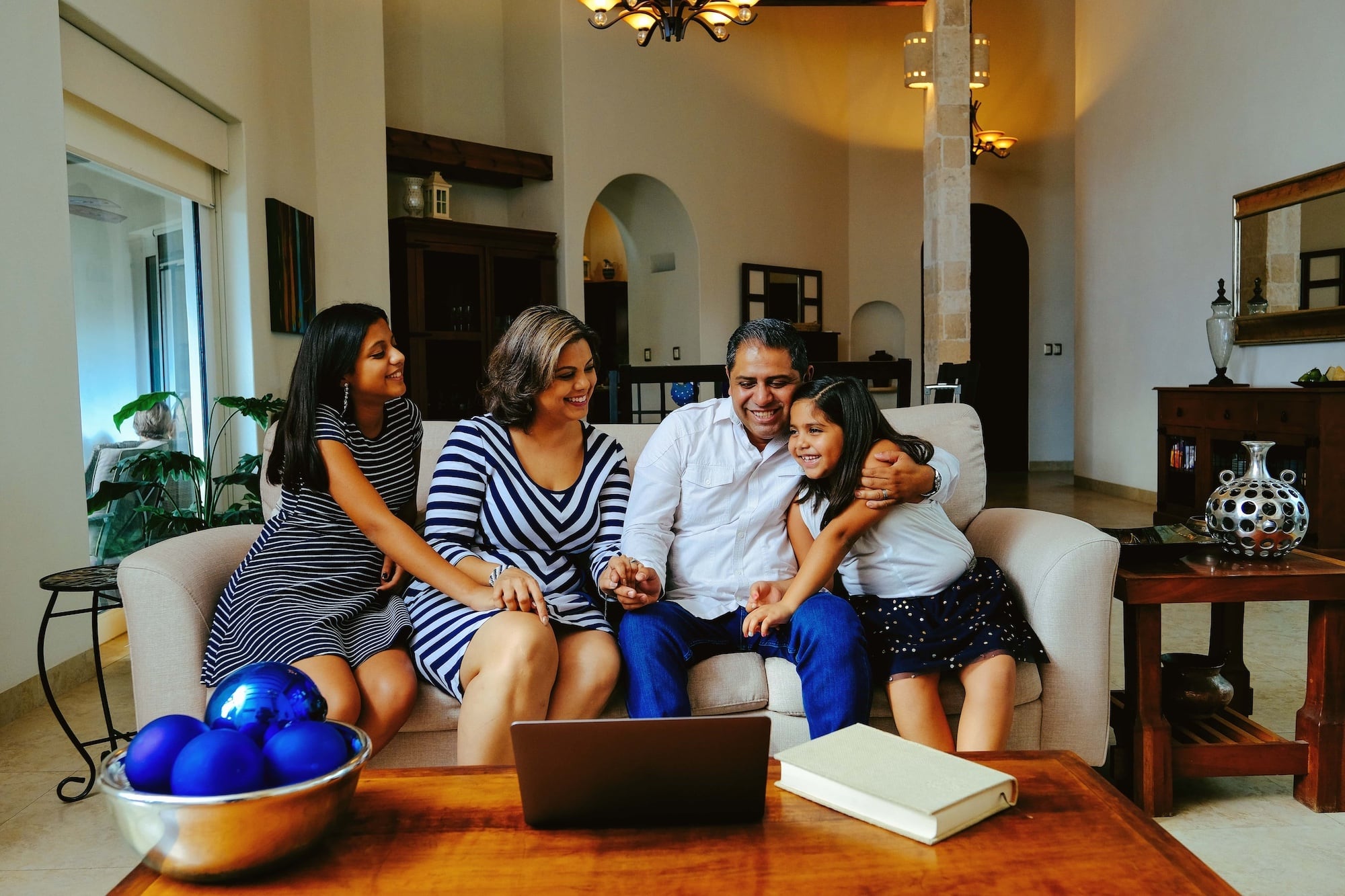 A family of four sits on a sofa in a warmly lit living room, smiling at a laptop on the table. The setting includes elegant decor with a vase of blue ornaments and a closed book. A woman and two young girls hug an older man.