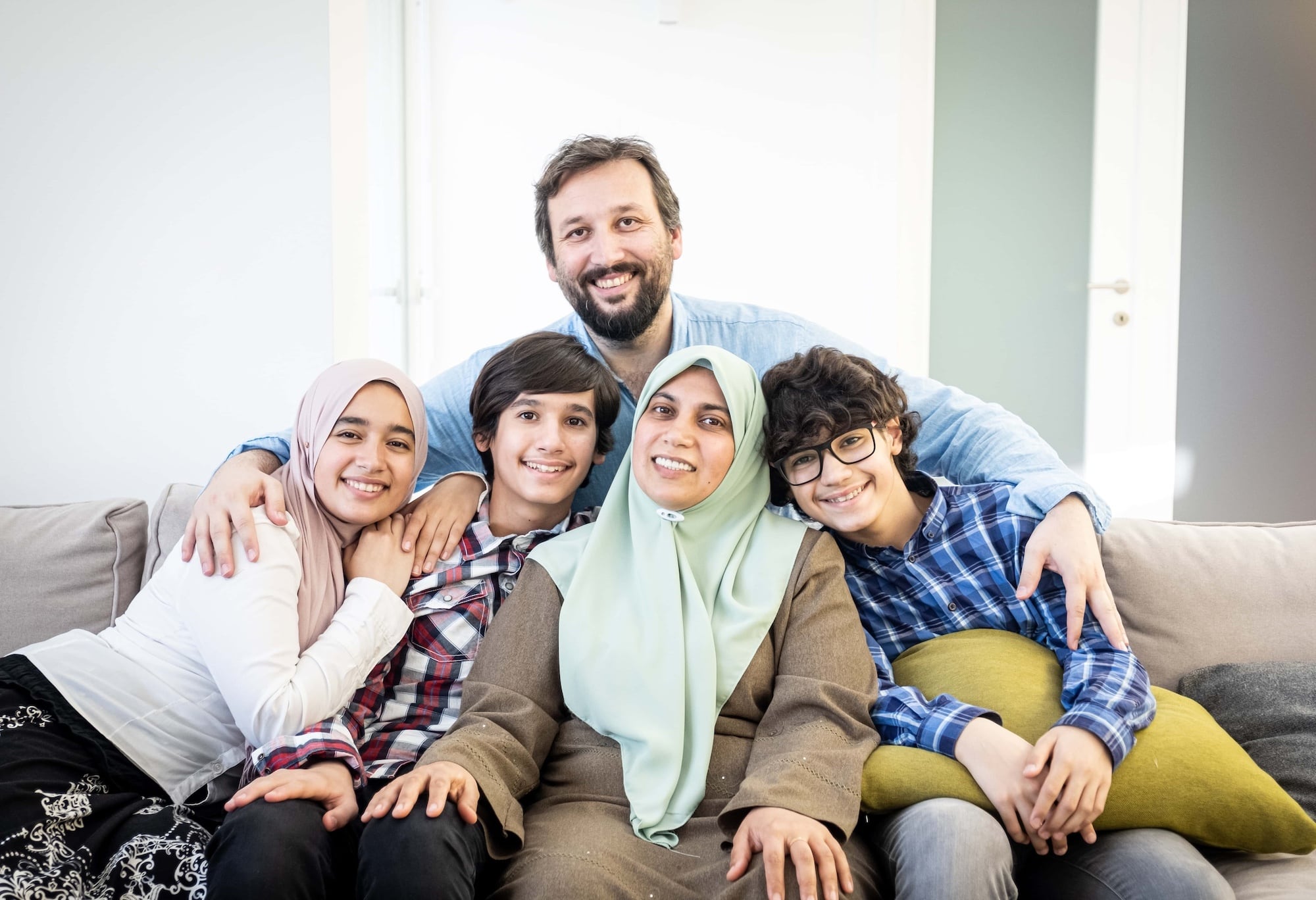 A family of five sits on a couch, smiling at the camera. The mother wears a green hijab, and the father has a beard. The children, two boys and a girl, are wearing plaid shirts. They appear joyful and relaxed in a bright room.