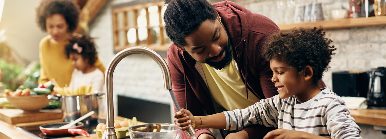 A man and child smile while washing dishes at a kitchen sink. In the background, a woman and another child prepare food on the countertop. The kitchen is warmly lit, with wooden shelves and a cozy atmosphere.