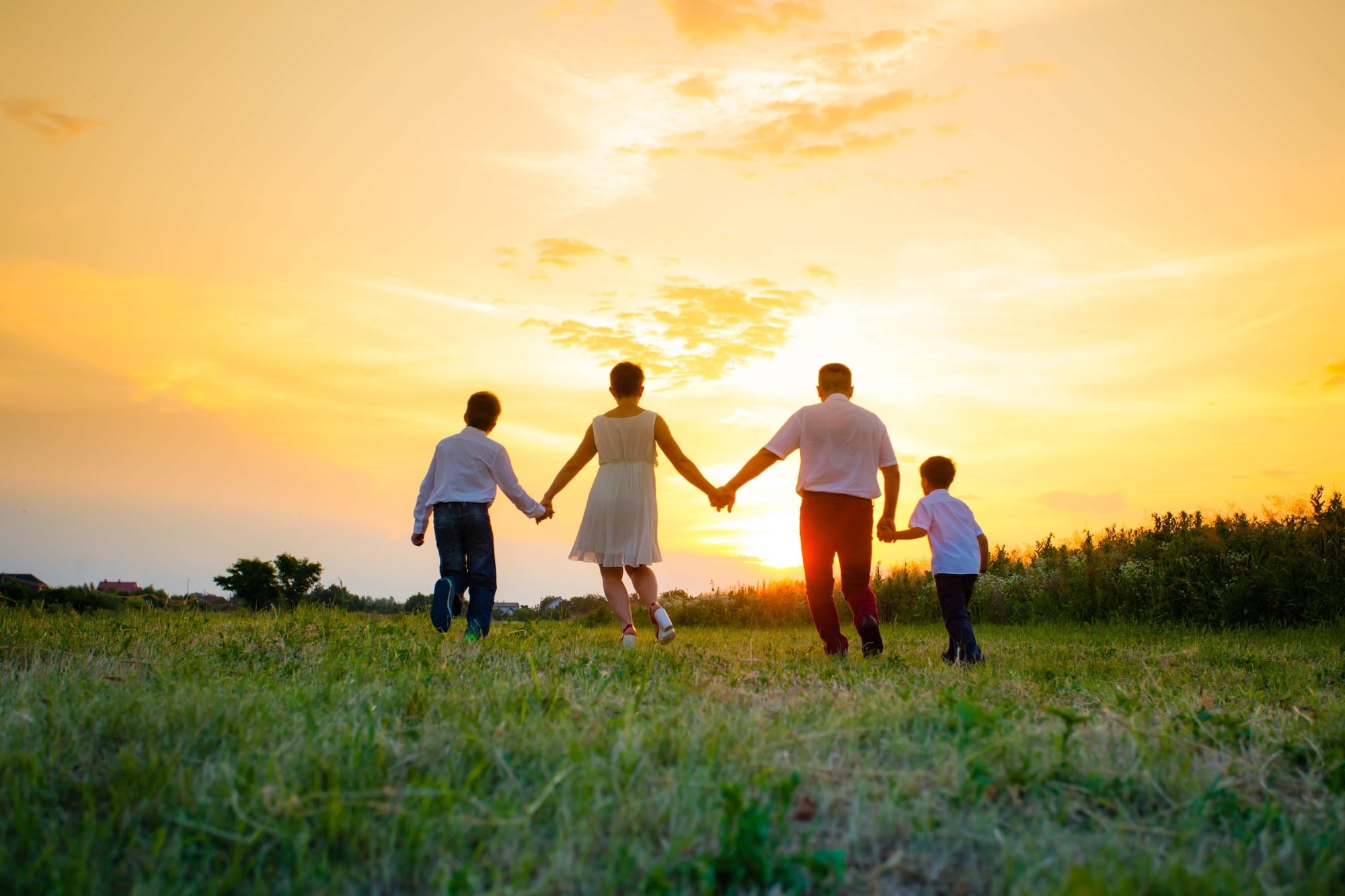Four people are walking hand in hand through a grassy field at sunset. The sky is a vibrant mix of orange and yellow hues. They are seen from behind, silhouetted against the colorful sky.