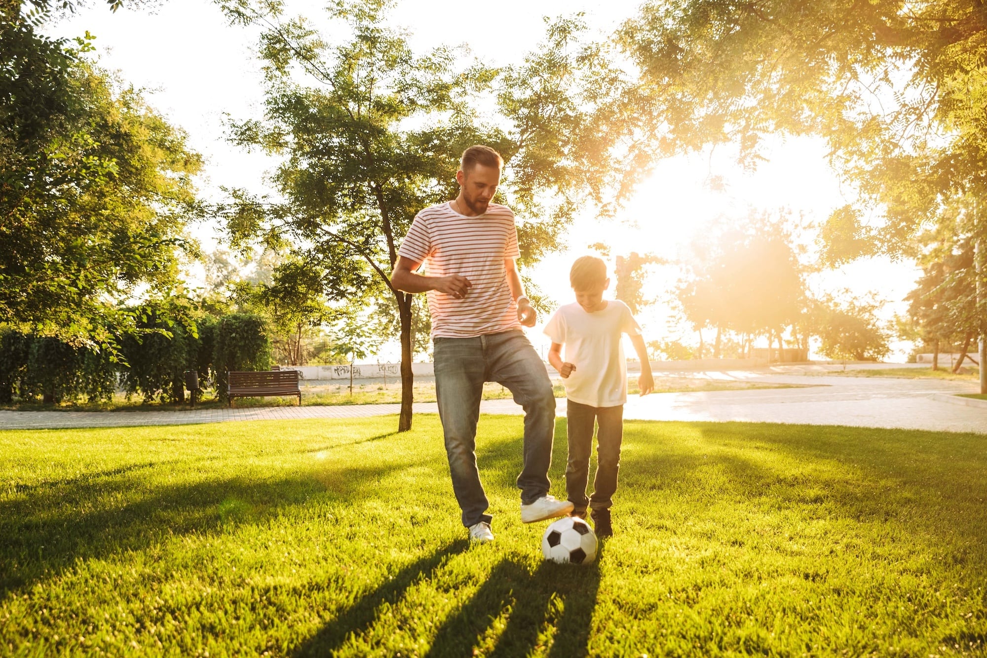 A man and a boy play with a soccer ball on a sunny day in a park. The man is in a striped shirt and jeans, and the boy is in a white shirt and jeans. Sunlight filters through the trees, casting long shadows on the grass.