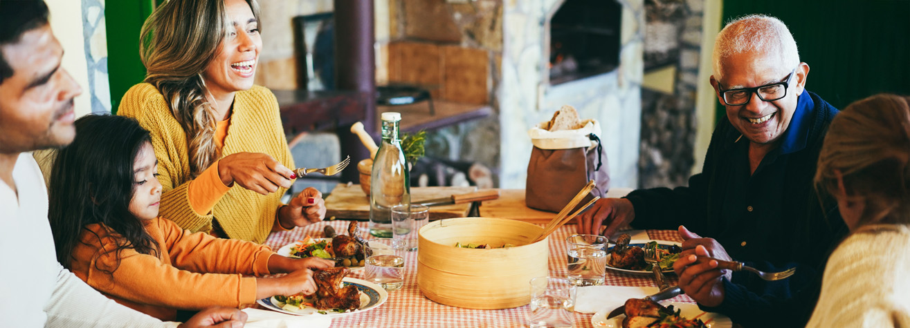 A joyful family shares a meal at a dining table. A young child, two adults, and two older adults are laughing and engaging with each other. Plates of food and drinks are spread around, enhancing the cozy, inviting atmosphere.