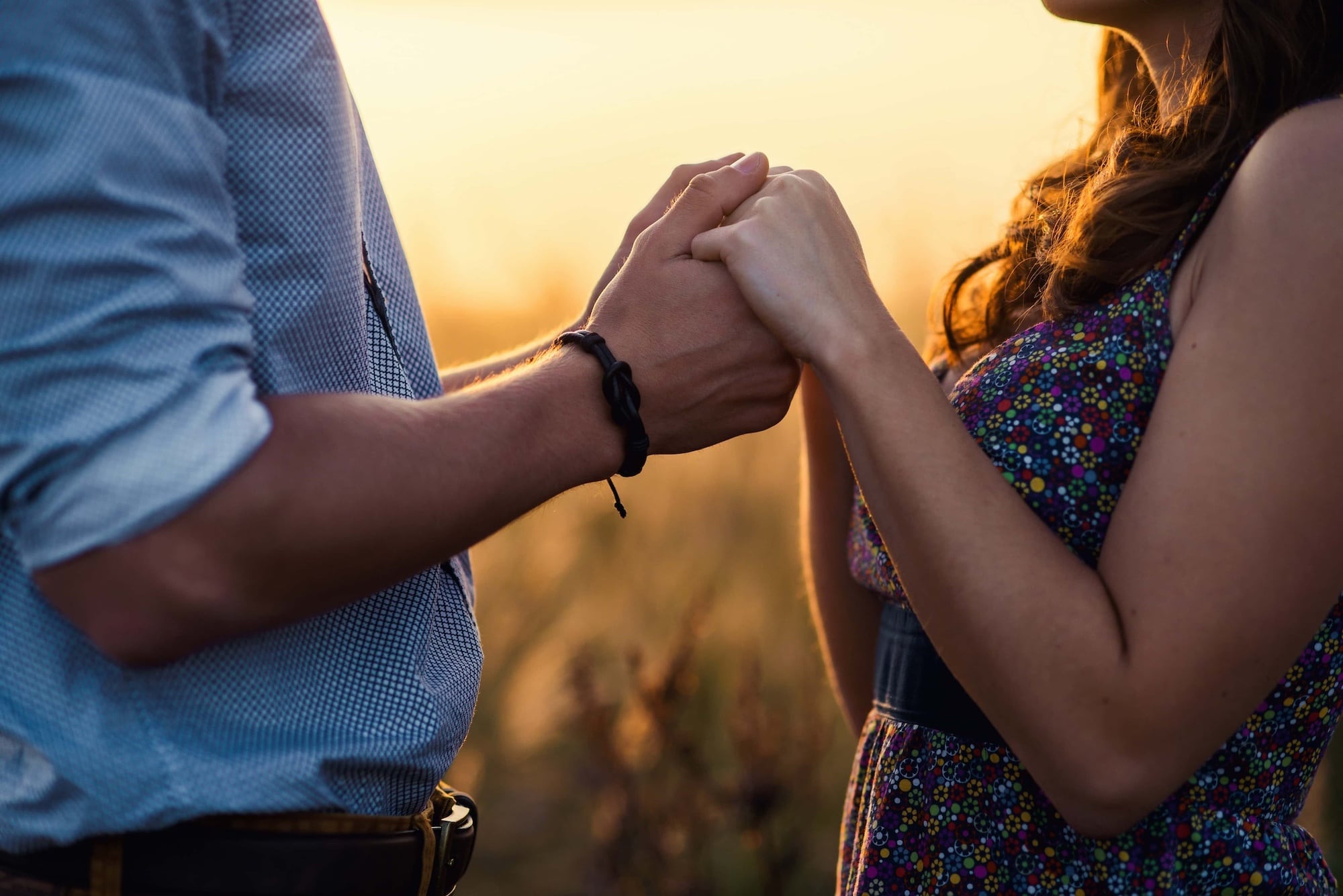 A couple holds hands at sunset, standing closely. The woman wears a floral dress, and the man wears a blue shirt with rolled-up sleeves. The warm, golden light creates a romantic atmosphere.