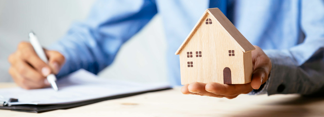 A person in a blue shirt holds a small wooden house model in one hand while signing documents with a pen on a clipboard with the other hand. The focus is on the house model, symbolizing real estate or mortgage concepts.