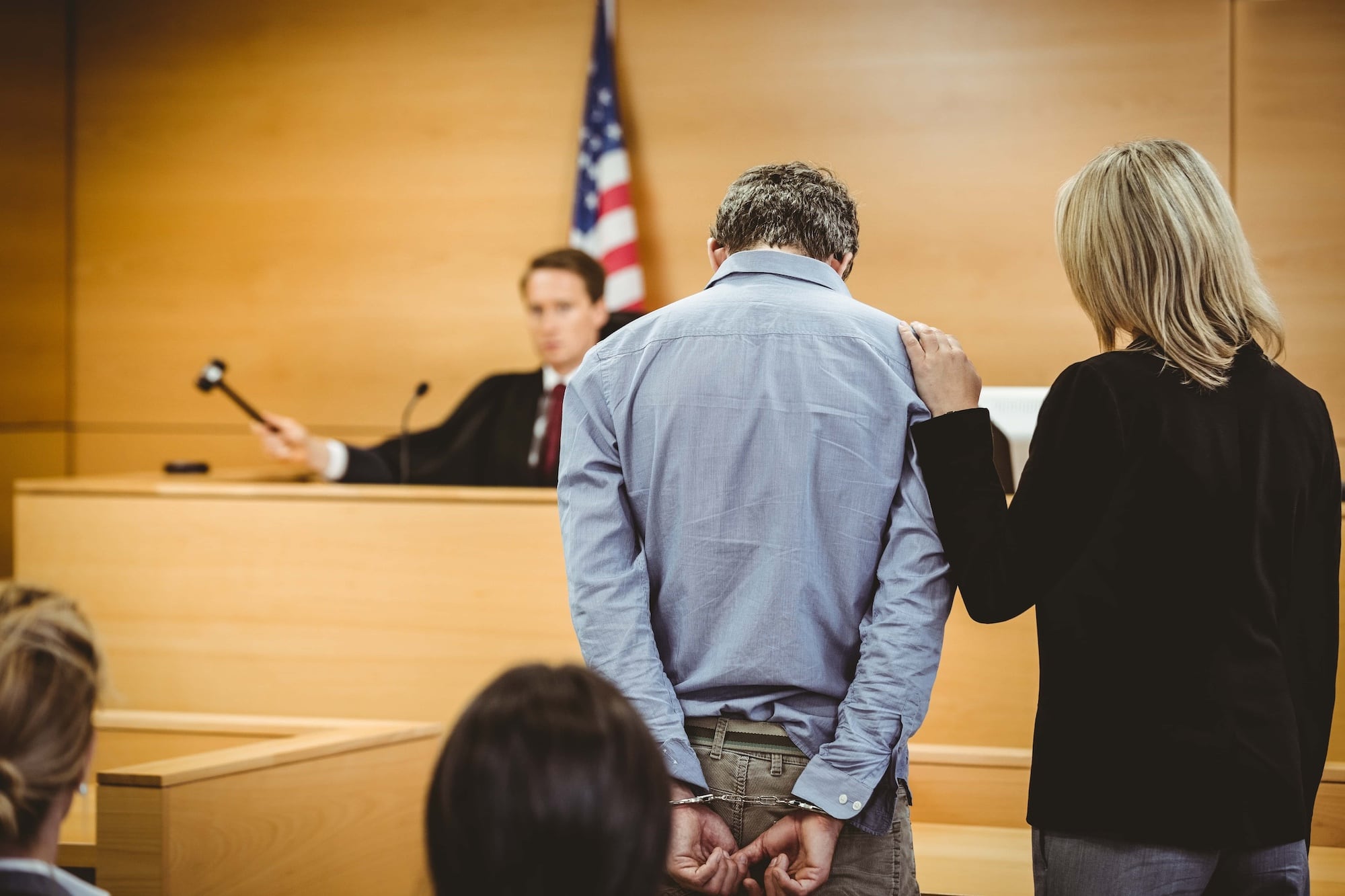 A man in handcuffs stands in a courtroom beside a woman with her hand on his shoulder. A judge is seated at a bench in the background, holding a gavel. An American flag is visible behind the judge.