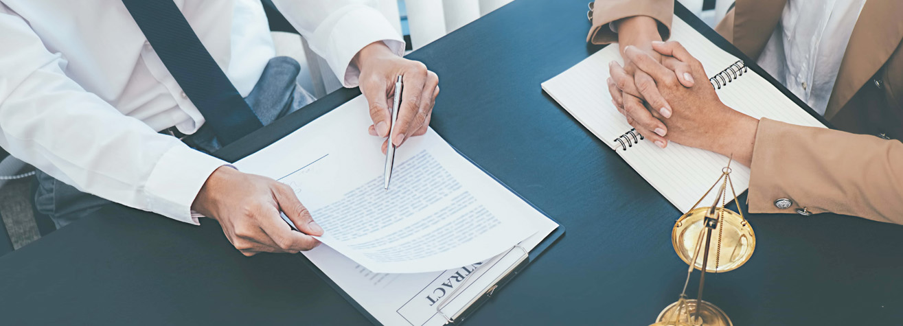 Two people at a table with a contract, pen, and notebook. One person is showing the document, while the other listens with hands clasped. A scale of justice is on the table, suggesting a legal or business meeting setting.