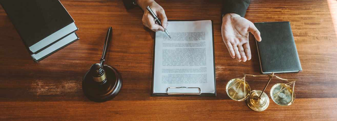 A person signing a document on a clipboard at a wooden desk. Nearby are a gavel, a stack of books, a black notebook, and a brass balance scale, suggesting a legal or judicial setting.
