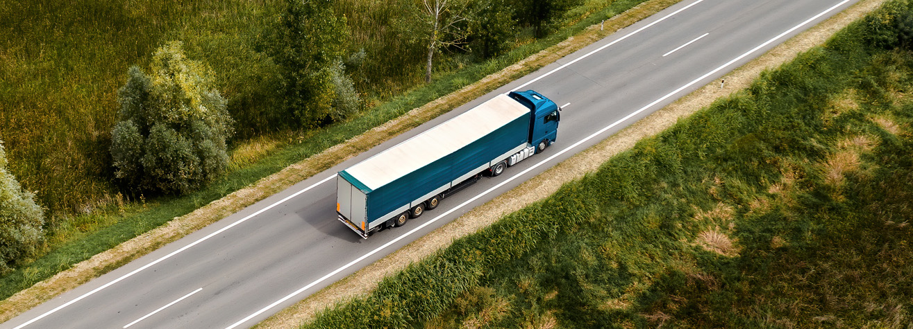Aerial view of a blue semi-truck with a white trailer driving on a straight rural road surrounded by green fields and trees. The road has two lanes, one going each direction. The landscape is mostly lush with some patches of brown grass.
