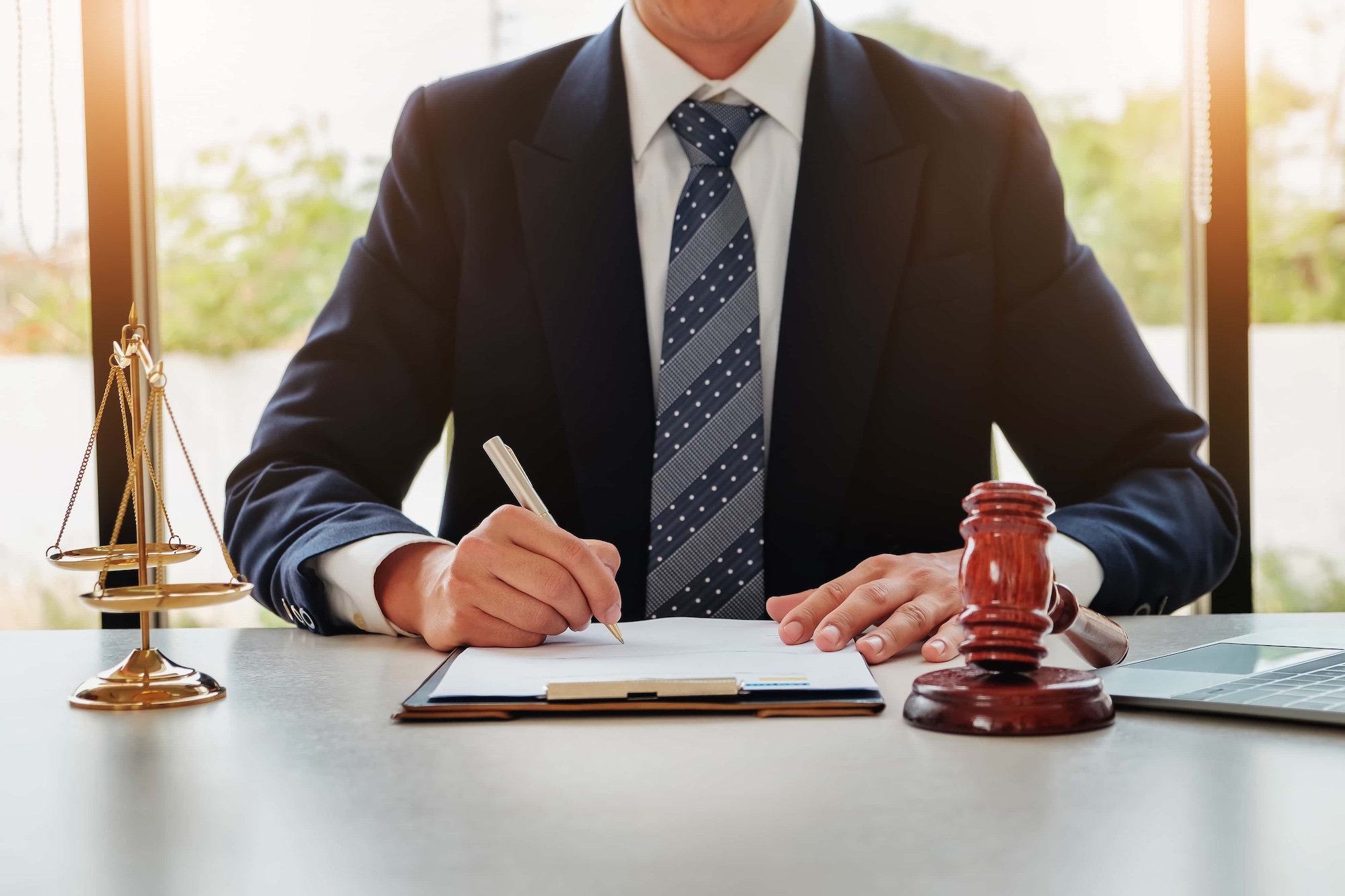 A person in a suit writes on paper at a desk with a pen. Nearby are a gavel, a set of scales, and a laptop. Bright lighting filters through a window in the background.