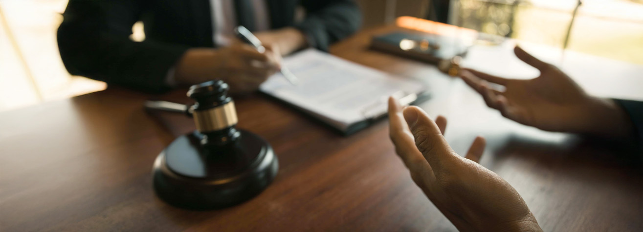 Two people sit at a wooden table discussing documents. One person gestures with hands visible, while the other writes on a clipboard. A judge's gavel is in the foreground, suggesting a legal or formal setting.